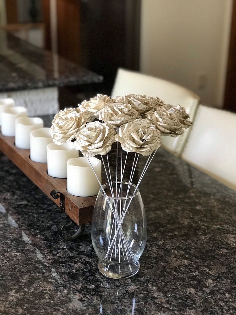 Puede incluir: Un jarr&oacute;n de cristal transparente contiene un ramo de rosas de papel. Las rosas est&aacute;n hechas de p&aacute;ginas de libro, con tallos de alambre blanco. Un portavelas de madera con velas blancas est&aacute; a la izquierda sobre una encimera de granito oscuro.