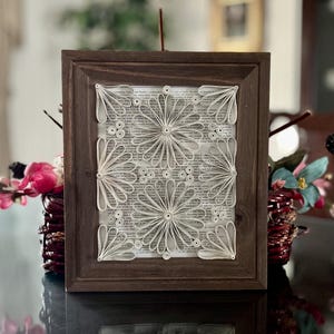 Puede incluir: Una obra de arte enmarcada que presenta un diseño de papel blanco sobre un fondo de página de libro. El diseño está hecho de tiras de papel enrolladas y crea un patrón geométrico de flores.