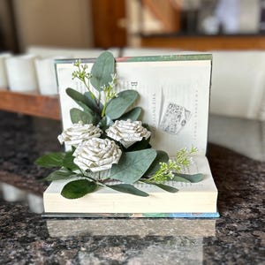 May include: An open book with three paper roses and greenery arranged on the pages. The roses are made from book pages, and the leaves are a mix of green and gray. The book is resting on a dark surface.