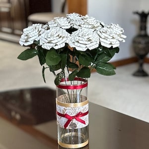 Puede incluir: Un ramo de rosas blancas de papel en un jarrón de vidrio transparente con una cinta de arpillera y encaje y un lazo rojo. El jarrón está sobre una mesa marrón oscura.