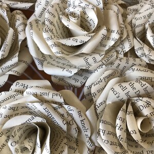 Puede incluir: Primer plano de varias rosas de papel hechas con p&aacute;ginas de libros. Las rosas son de color crema con texto visible de las p&aacute;ginas del libro. Los p&eacute;talos est&aacute;n cuidadosamente rizados para parecerse a rosas reales, creando un arreglo floral &uacute;nico.