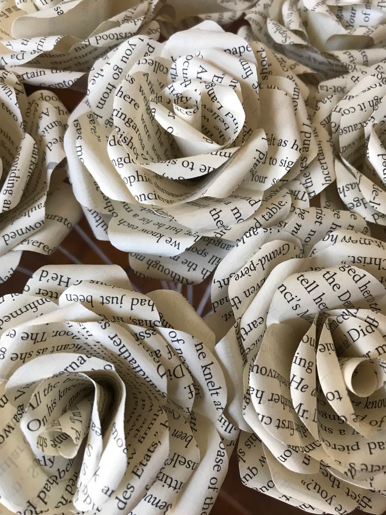 Puede incluir: Primer plano de varias rosas de papel hechas con p&aacute;ginas de libros. Las rosas son de color crema con texto visible de las p&aacute;ginas del libro. Los p&eacute;talos est&aacute;n cuidadosamente rizados para parecerse a rosas reales, creando un arreglo floral &uacute;nico.