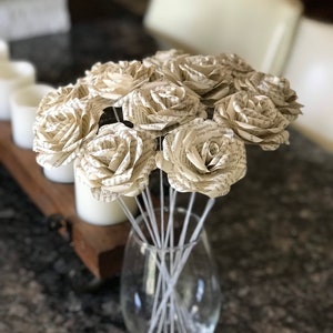 Puede incluir: Un ramo de rosas de papel hechas con p&aacute;ginas de libros. Las flores de color crema est&aacute;n dispuestas en un jarr&oacute;n de cristal transparente con tallos blancos. Las rosas est&aacute;n en plena floraci&oacute;n, con texto visible.