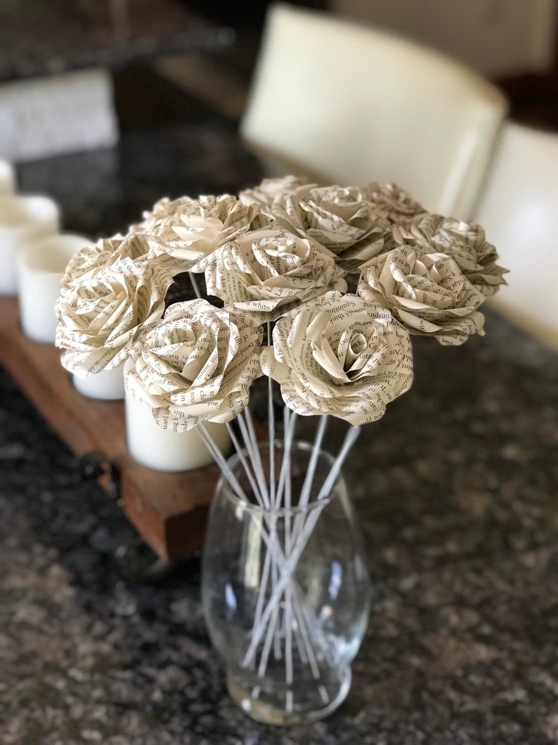 Puede incluir: Un ramo de rosas de papel hechas con p&aacute;ginas de libros. Las flores de color crema est&aacute;n dispuestas en un jarr&oacute;n de cristal transparente con tallos blancos. Las rosas est&aacute;n en plena floraci&oacute;n, con texto visible.
