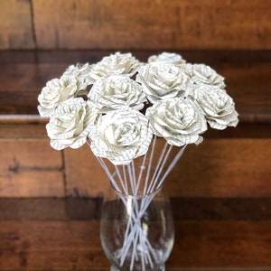 Puede incluir: Un ramo de rosas de papel hechas con p&aacute;ginas de libros, exhibido en un jarr&oacute;n de vidrio transparente. Las rosas son de color crema con texto visible, y los tallos son blancos. El fondo es una superficie de madera.