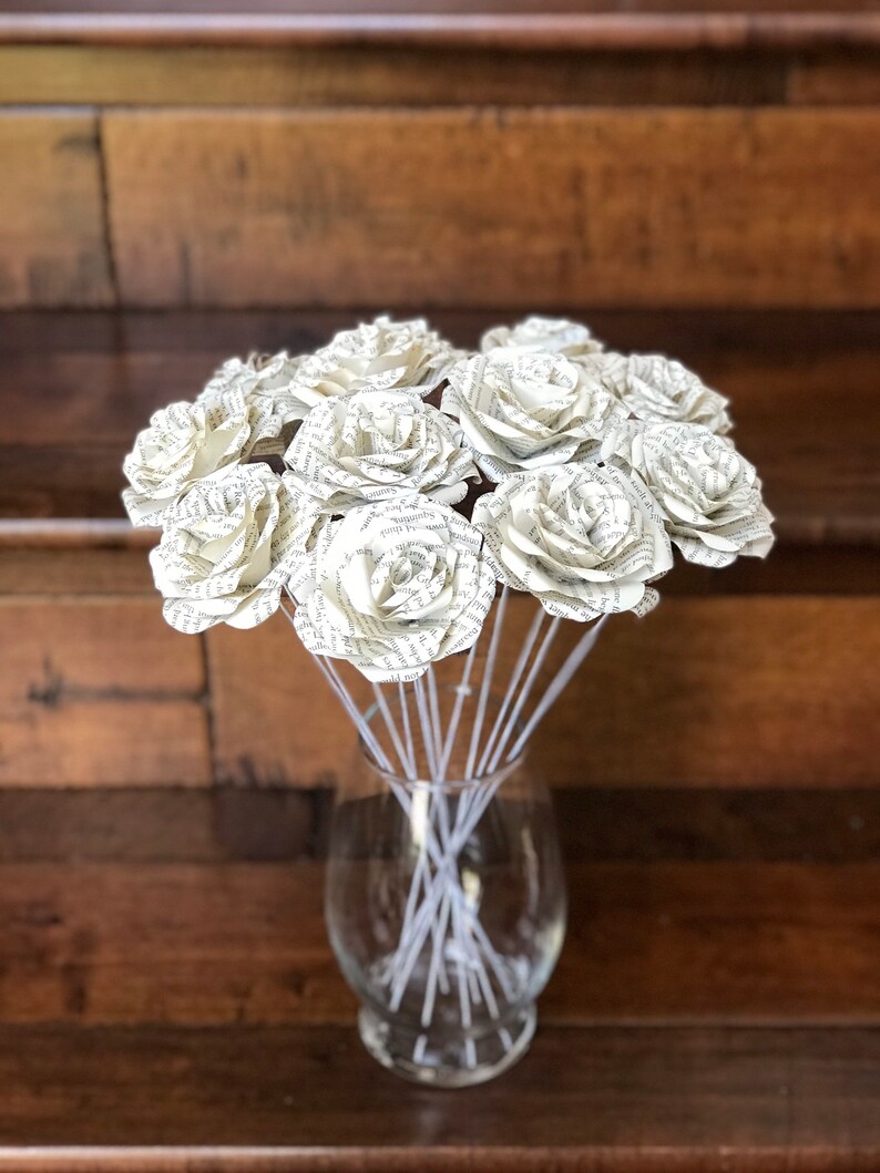 Puede incluir: Un ramo de rosas de papel hechas con p&aacute;ginas de libros, exhibido en un jarr&oacute;n de vidrio transparente. Las rosas son de color crema con texto visible, y los tallos son blancos. El fondo es una superficie de madera.