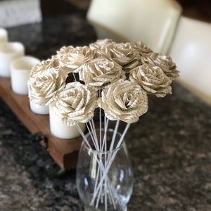 Puede incluir: Un ramo de rosas de papel hechas con p&aacute;ginas de libros en un jarr&oacute;n de cristal transparente. Las rosas son de color crema con texto visible, y el jarr&oacute;n est&aacute; sobre una encimera oscura. El arreglo incluye m&uacute;ltiples tallos, creando una exhibici&oacute;n floral completa.