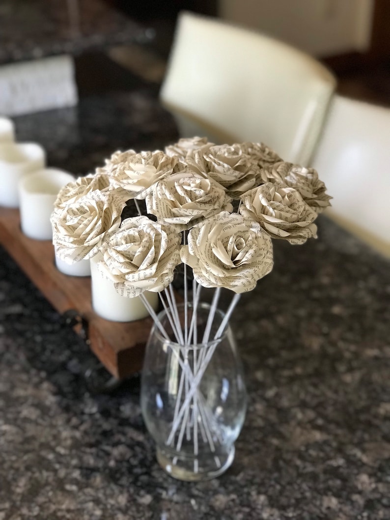 Puede incluir: Un ramo de rosas de papel hechas con p&aacute;ginas de libros en un jarr&oacute;n de cristal transparente. Las rosas son de color crema con texto visible, y el jarr&oacute;n est&aacute; sobre una encimera oscura. El arreglo incluye m&uacute;ltiples tallos, creando una exhibici&oacute;n floral completa.