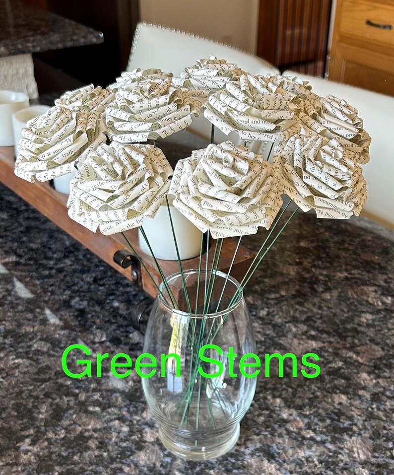 Puede incluir: Un ramo de rosas de papel hechas con p&aacute;ginas de libros, exhibido en un jarr&oacute;n de vidrio transparente. Las rosas tienen tallos de alambre verde. El texto "Green Stems" est&aacute; escrito en verde.