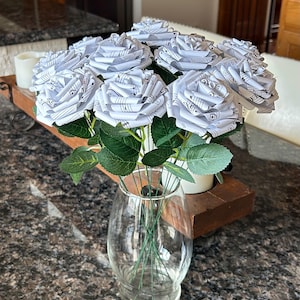 Puede incluir: Un ramo de rosas de papel hechas con partituras, exhibido en un jarrón de cristal transparente. Las rosas blancas tienen hojas y tallos verdes. El jarrón se encuentra sobre una bandeja de madera marrón oscuro con una vela blanca.