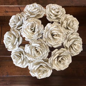 Puede incluir: Una corona circular compuesta por rosas de papel. Cada rosa est&aacute; hecha de p&aacute;ginas de un libro, mostrando texto visible. Las rosas est&aacute;n dispuestas en un c&iacute;rculo completo, creando un elemento decorativo &uacute;nico. Fondo de madera oscura.