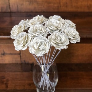 Puede incluir: Un ramo de rosas de papel hechas con p&aacute;ginas de libros, exhibido en un jarr&oacute;n de vidrio transparente. Las rosas son blancas con texto visible, y los tallos son finos y blancos. El jarr&oacute;n se encuentra sobre un fondo de madera oscura.