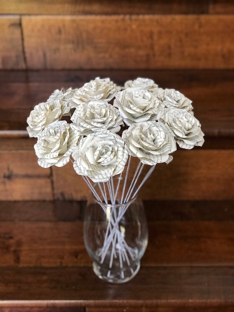 Puede incluir: Un ramo de rosas de papel hechas con p&aacute;ginas de libros, exhibido en un jarr&oacute;n de vidrio transparente. Las rosas son blancas con texto visible, y los tallos son finos y blancos. El jarr&oacute;n se encuentra sobre un fondo de madera oscura.