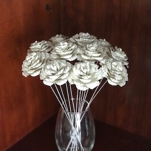 May include: A bouquet of white paper roses made from book pages in a clear glass vase. The roses are arranged in a circular pattern and the stems are white.