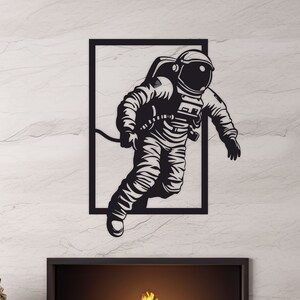 Peut inclure: Décoration murale en métal noir représentant la silhouette d'un astronaute flottant dans l'espace. L'astronaute porte une combinaison spatiale et un casque. L'œuvre est encadrée dans un rectangle noir.