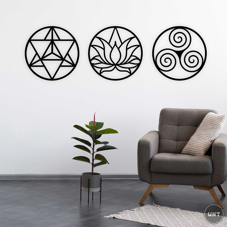 May include: Three black metal wall decor pieces in the shape of circles. The circles feature geometric designs, including a lotus flower, a triskelion, and a geometric star. The decor is displayed on a white wall with a gray armchair and a potted plant in the foreground.