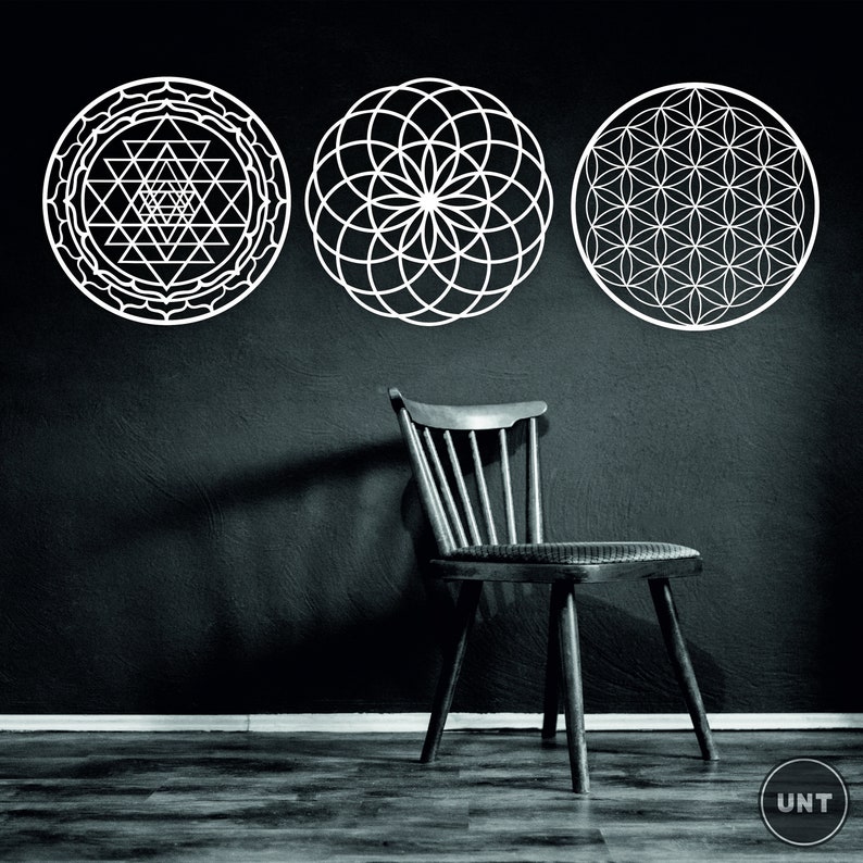May include: Three white geometric designs on a black wall. The designs are a flower of life, a sri yantra, and a seed of life. A wooden chair sits in front of the designs.