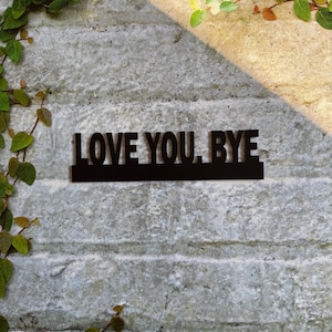 Könnte beinhalten: Schwarzes Holzschild mit dem Text "LOVE YOU, BYE" an einer grauen Backsteinwand mit grünen Ranken.