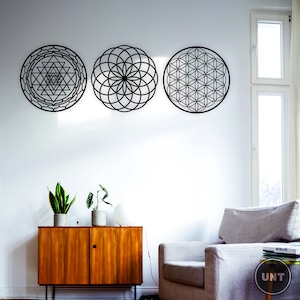 May include: Three black metal wall decor pieces with geometric designs. The designs are a flower of life, a sri yantra, and a seed of life. The decor is hanging on a white wall in a living room setting.