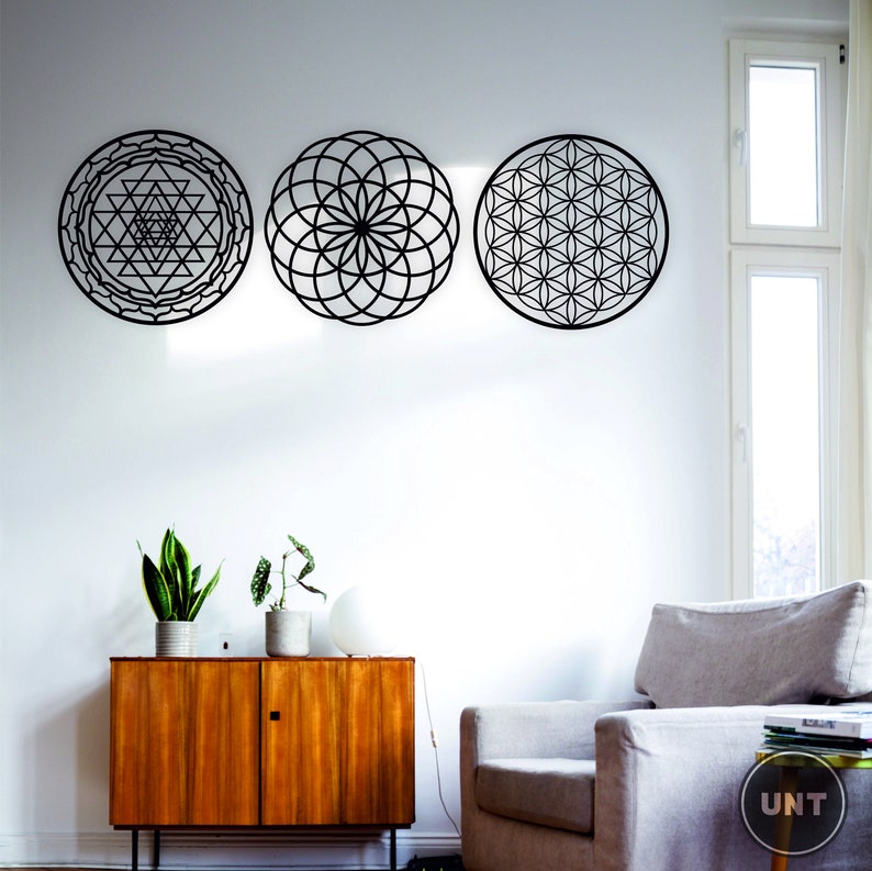 May include: Three black metal wall decor pieces with geometric designs. The designs are a flower of life, a sri yantra, and a seed of life. The decor is hanging on a white wall in a living room setting.