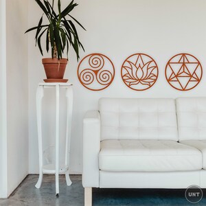 May include: Three circular wall decor pieces in a light brown color, featuring a stylized triquetra, a lotus flower, and a geometric star. The decor is mounted on a white wall above a white couch.