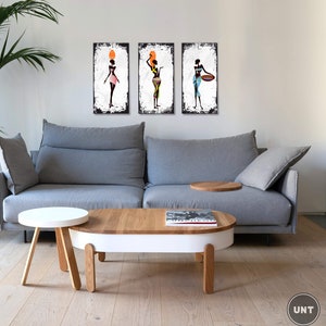 May include: Three framed canvas prints of stylized black women in colorful clothing. The women are holding various objects, including a pot, a bowl, and a basket. The prints are displayed on a white wall above a gray sofa and a wooden coffee table.