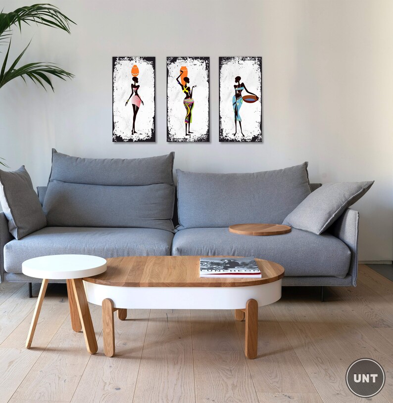 May include: Three framed canvas prints of stylized black women in colorful clothing. The women are holding various objects, including a pot, a bowl, and a basket. The prints are displayed on a white wall above a gray sofa and a wooden coffee table.