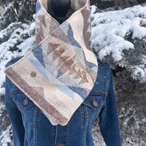 May include: A patterned cowl scarf in shades of beige, blue, and brown, secured with three brass-colored buttons. The scarf is displayed on a black mannequin head over a denim jacket, with a snowy background.