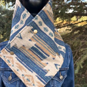 Pode incluir: Um aquecedor de pescoço com padrão azul, bege e branco com botões cor de latão. O aquecedor de pescoço é feito de um tecido grosso e tecido e é usado sobre uma jaqueta jeans. O design inclui listras e formas geométricas.