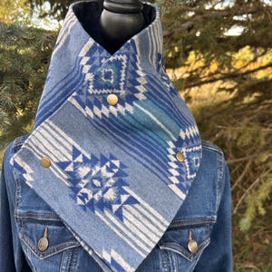 May include: A blue and white patterned cowl neck warmer with gold-colored buttons. The cowl is layered over a denim jacket. The pattern includes geometric shapes and stripes in shades of blue and white.