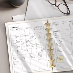 May include: Open planner with a January 2026 calendar, gold binder rings, and lined pages for notes. The planner includes sections for goals, priorities, and to-do lists. A pair of eyeglasses and a ceramic mug are visible.