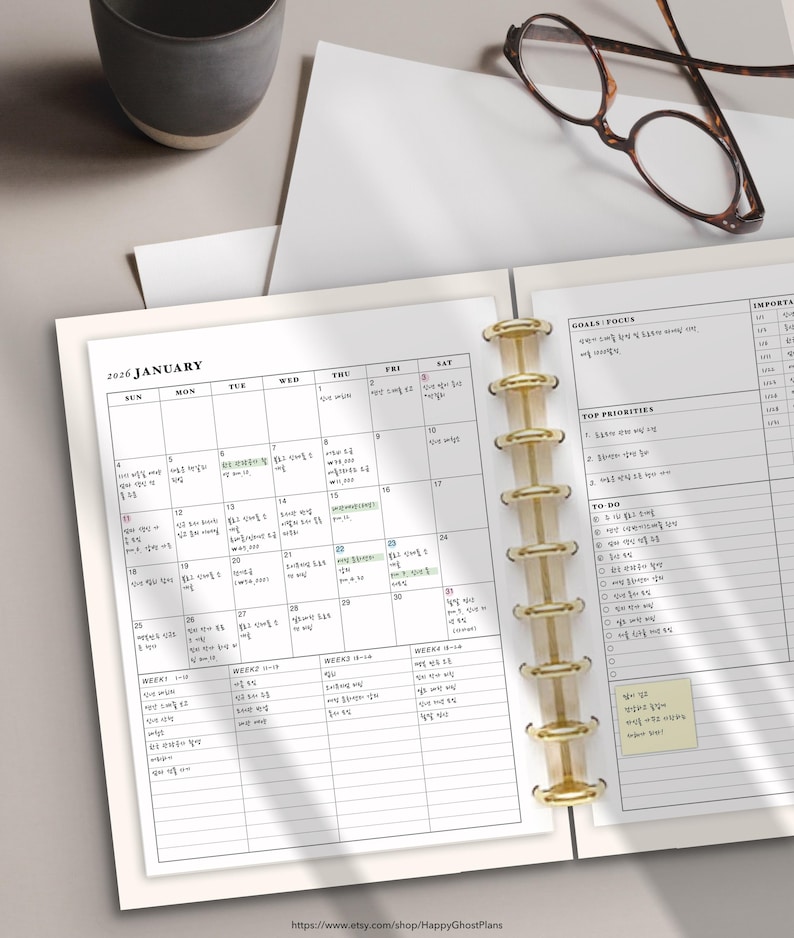 May include: Open planner with a January 2026 calendar, gold binder rings, and lined pages for notes. The planner includes sections for goals, priorities, and to-do lists. A pair of eyeglasses and a ceramic mug are visible.