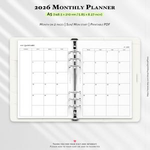May include: A white A5 2026 monthly planner, open to a January calendar page. The planner has a binder mechanism and includes the text "2026 Monthly Planner" and "A5 (14.85 x 21.0 cm)". The calendar is designed to be printable.