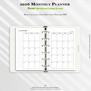 May include: A white 2026 monthly planner, pocket-sized at 8.1 x 12 cm. The planner displays a January calendar layout. Text on the planner reads "Month on 2 pages | Sun/Mon start | Printable PDF". The planner is held open by a silver binder.