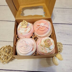 May include: A gift box containing four baby items shaped like cupcakes. The cupcakes are pink and white, with decorative bows and wooden cupcake toppers. The box is brown with a clear window, and the cupcakes sit on shredded paper.