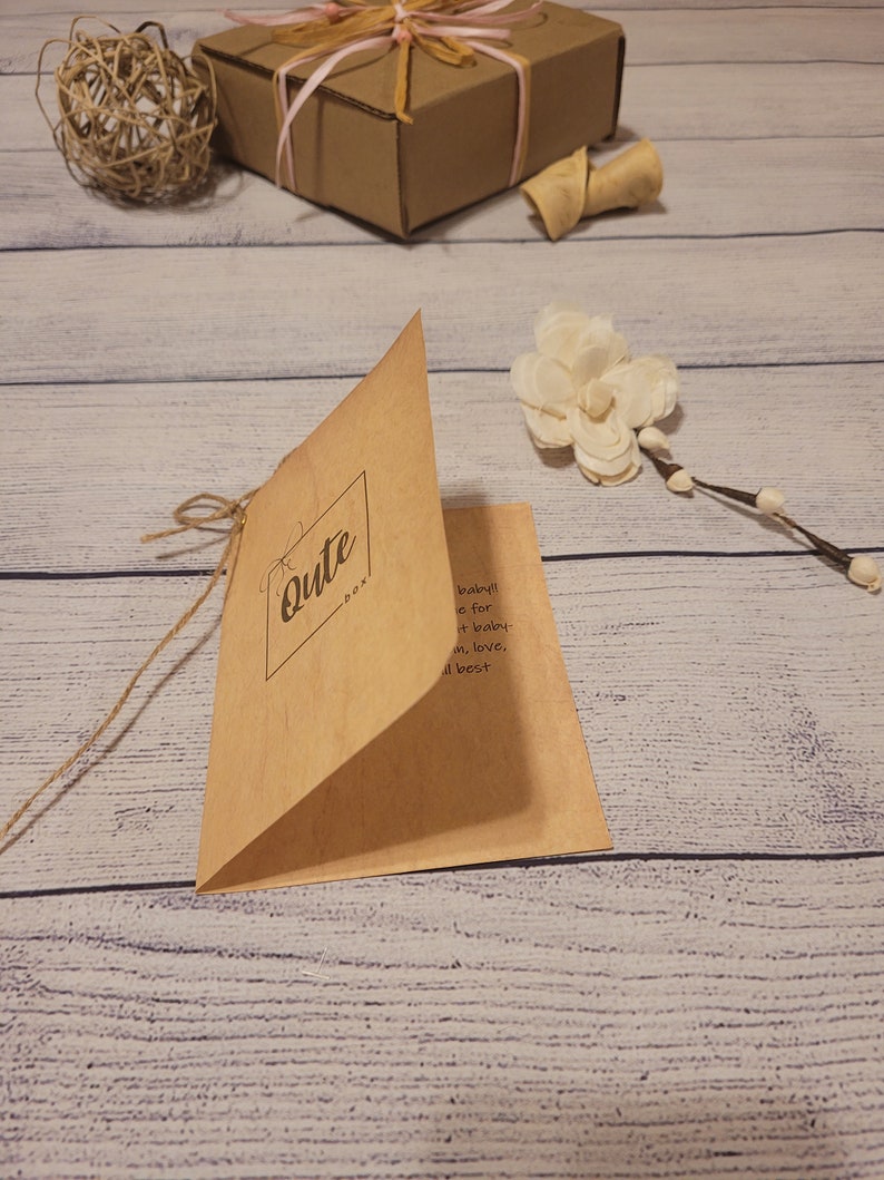 May include: A kraft paper card with the word "Cute" in a square frame, tied with twine. A brown gift box with pink and beige ribbon, a decorative ball, and a white flower are also visible.