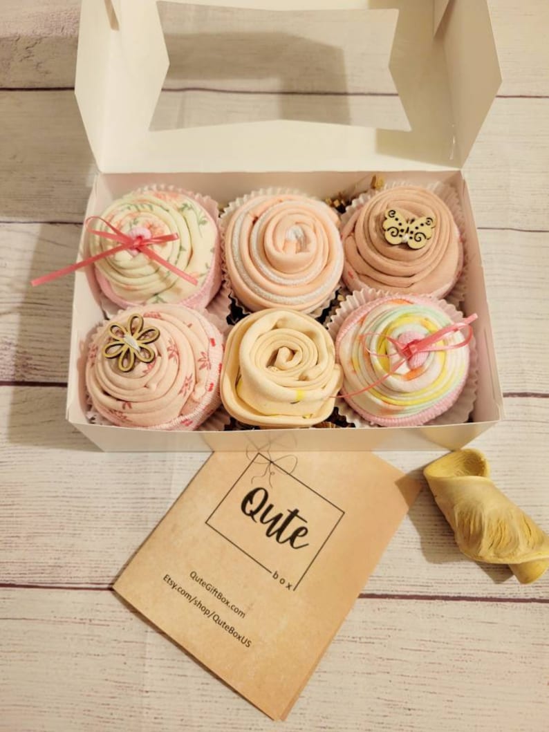 May include: A white box containing six decorative items resembling cupcakes. Each is a different color and design, with some featuring wooden embellishments and ribbons. A small card with the word "Qute" is visible.