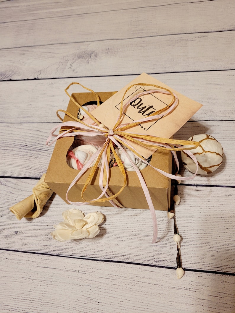 May include: A brown gift box with four compartments, tied with pink and natural raffia ribbon. The box contains small, wrapped items. A small card with the word "Qute" is attached. Decorative elements include a white flower and a decorative ball.