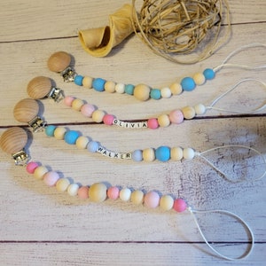 May include: Three pacifier clips with wooden beads in pastel colors, including pink, blue, and cream. Two clips have names spelled out in letter beads: "OLIVIA" and "WALKER". Each clip has a wooden clip and a white cord.
