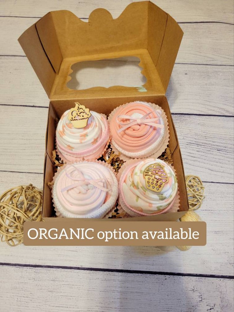 May include: A gift box with four cupcake-shaped baby socks. The socks are pink and white, decorated with ribbons and wooden cupcake toppers. The box is brown with a clear window. Text on the image reads "ORGANIC option available."