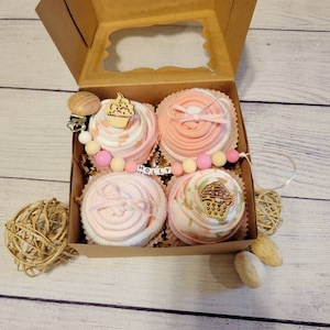 May include: A gift box with four baby items arranged to resemble cupcakes. Each 'cupcake' is pink and white, decorated with wooden cupcake toppers and ribbons. The box includes a pacifier clip and the name "HOLLY".