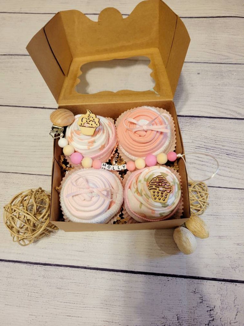 May include: A gift box with four baby items arranged to resemble cupcakes. Each 'cupcake' is pink and white, decorated with wooden cupcake toppers and ribbons. The box includes a pacifier clip and the name "HOLLY".