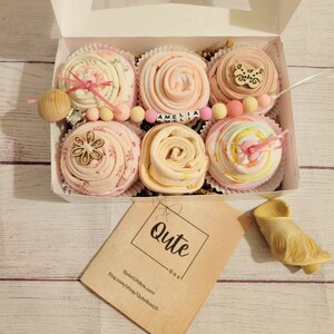 May include: A white box containing six baby gifts, each resembling a cupcake. The gifts are made of pink and white fabric, with decorative elements like wooden beads and a pacifier. The box also includes a tag that says "Qute box".