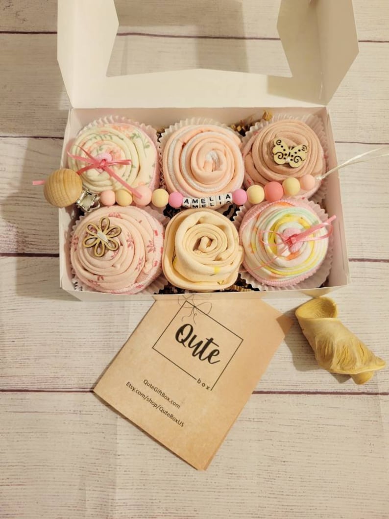 May include: A white box containing six baby gifts, each resembling a cupcake. The gifts are made of pink and white fabric, with decorative elements like wooden beads and a pacifier. The box also includes a tag that says "Qute box".