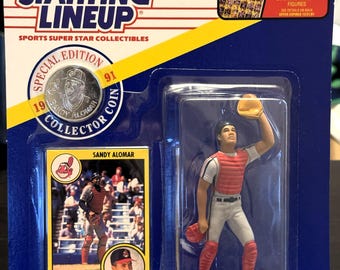 May include: A Starting Lineup sports collectible featuring a baseball player figurine in a red and white uniform, a baseball card, and a collector coin. The packaging includes the text "Starting Lineup" and "Sandy Alomar".