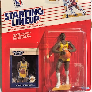 Puede incluir: Una figura coleccionable de Starting Lineup de Magic Johnson con uniforme de los Lakers, sosteniendo un balón de baloncesto. El embalaje incluye una tarjeta comercial y las palabras "Starting Lineup" y "NBA Official Licensed Product". La figura está en una funda de plástico transparente.