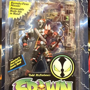 May include: Commando Spawn action figure in a plastic package. The figure is black and red, with a removable knife and bazooka missile. The packaging includes the text "America's Best Selling Comic Book."