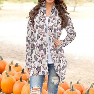 May include: A white cardigan with a long-sleeve design featuring a pattern of colorful books. The cardigan is open in the front and has a relaxed fit.