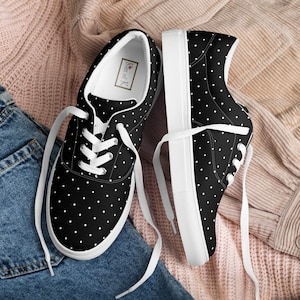 Puede incluir: Un par de zapatillas bajas de lona negras con lunares blancos y cordones blancos. Las zapatillas tienen una suela de goma blanca y están sobre un fondo de tela rosa y azul. El nombre de la marca "Kiss of K" está impreso en el interior de la zapatilla.