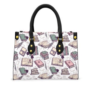 May include: A white handbag with a black handle and gold hardware. The bag is covered in a pattern of books, pens, and ink splatters in shades of green, pink, and beige. The bag has a rectangular shape.